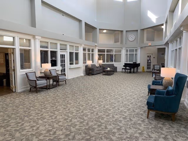A spacious and well-lit common area in a senior living facility with high ceilings and large windows. The room features multiple seating arrangements including armchairs and sofas with side tables and lamps. A black grand piano is positioned near the back wall under a large clock. The walls have multiple windows and doors leading to other rooms.