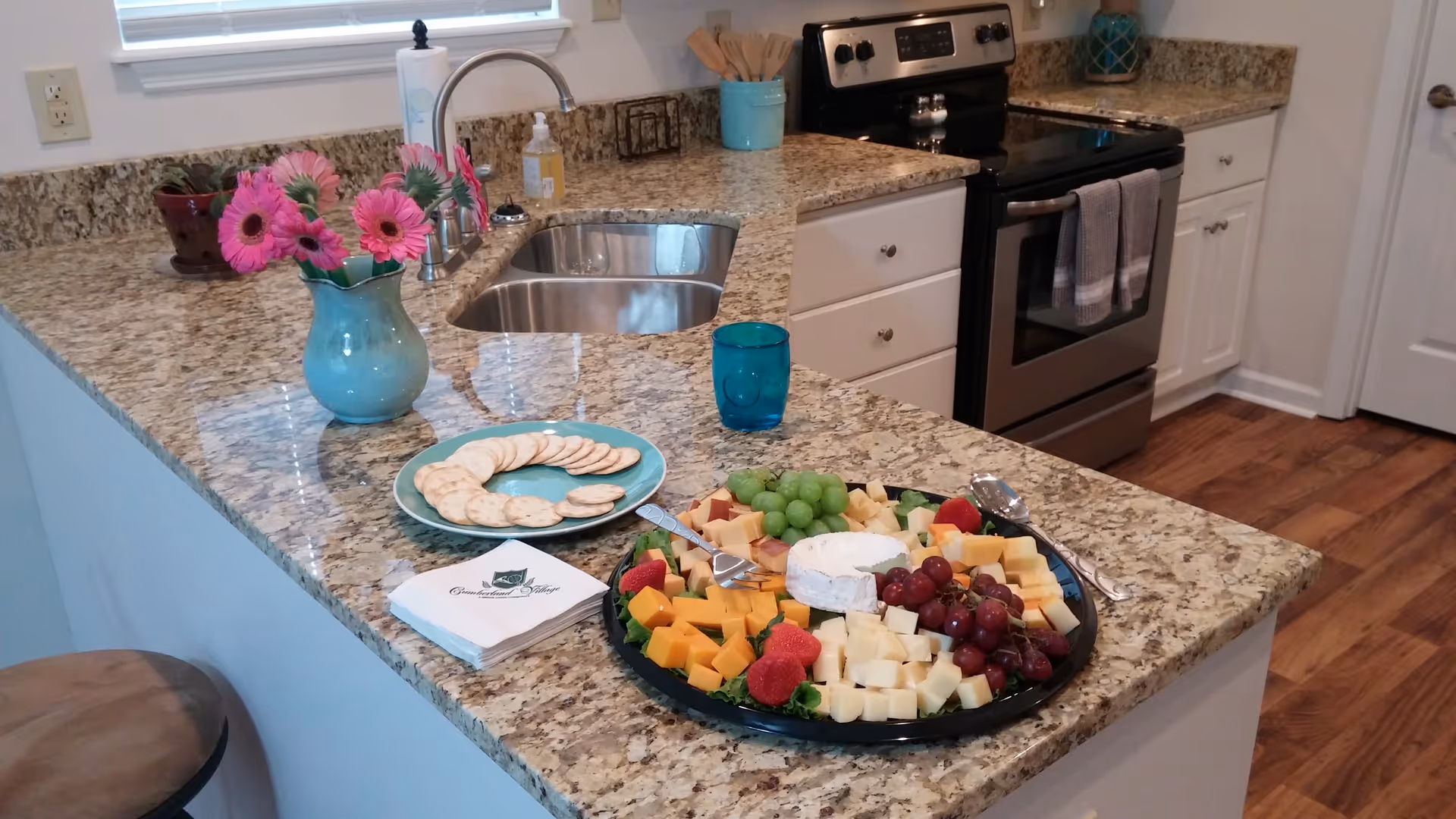 Kitchen island with a granite countertop set with a cheese and fruit platter, crackers, a vase of pink flowers, sink and stove in the background.