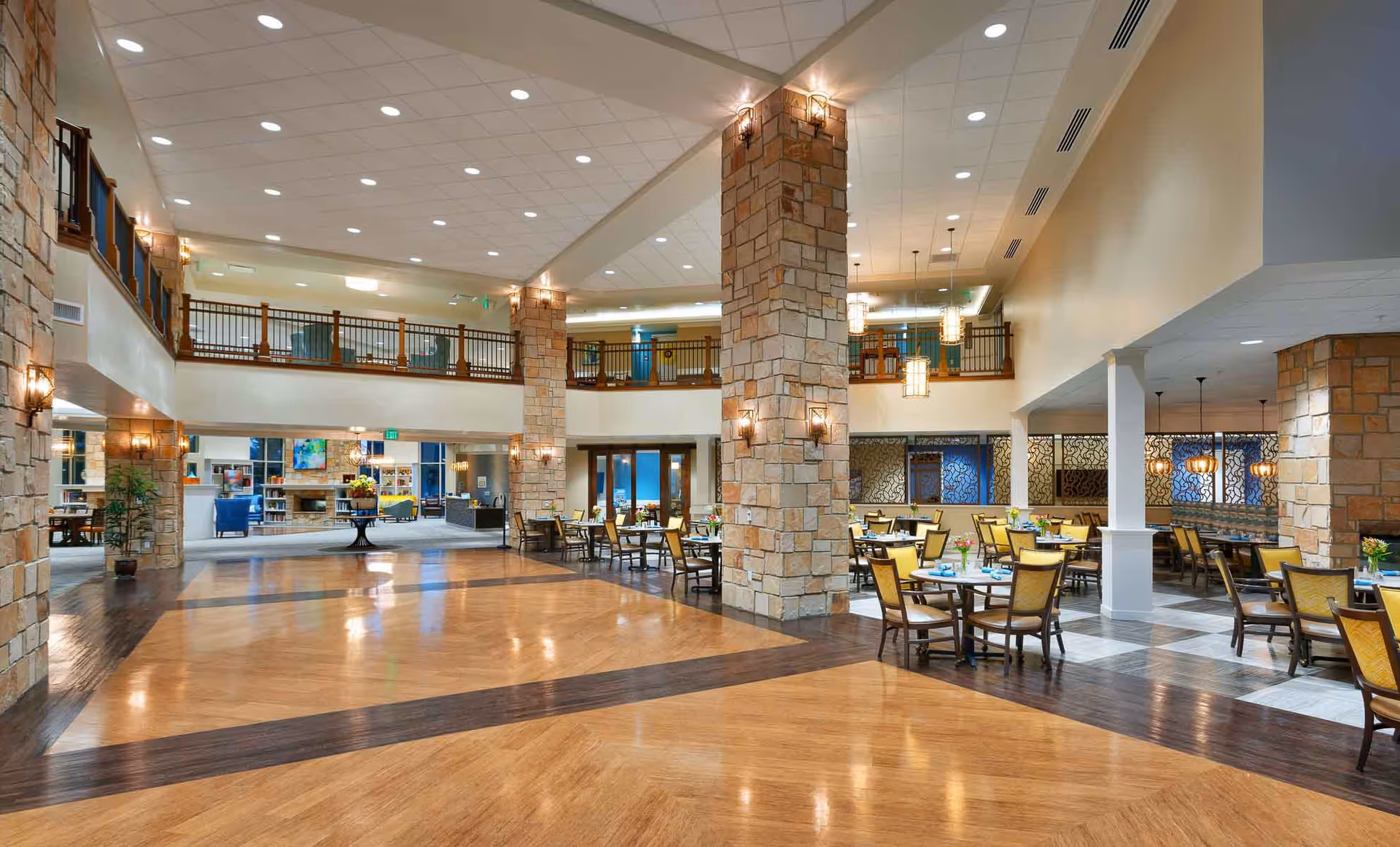 Spacious and well-lit dining area in a senior living facility with multiple tables and chairs arranged neatly. The room features stone pillars, a high ceiling with recessed lighting, and a second-floor balcony overlooking the space. There is a cozy seating area with armchairs and a fireplace visible in the background.
