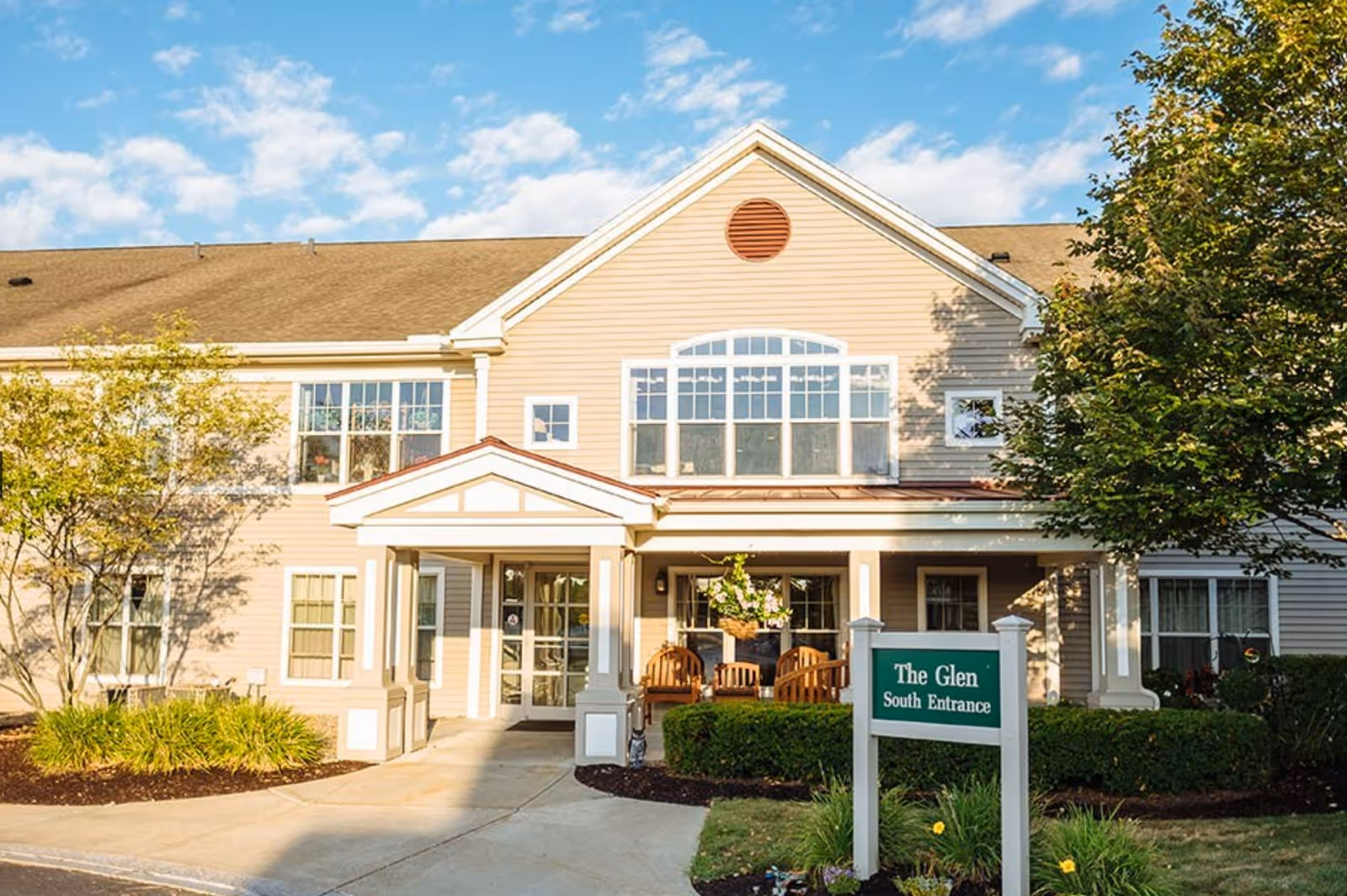 Exterior view of The Glen at Hiland Meadows showing the south entrance with a covered porch, wooden chairs, and surrounding greenery under a partly cloudy blue sky.