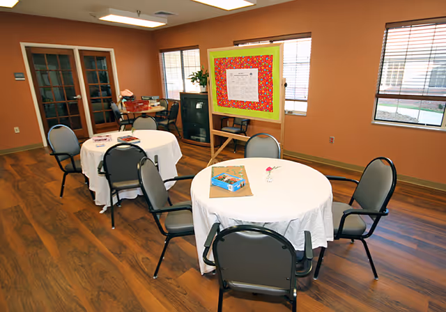 A bright communal room with round tables draped in white cloths, chairs, windows, and a bulletin board on an easel.