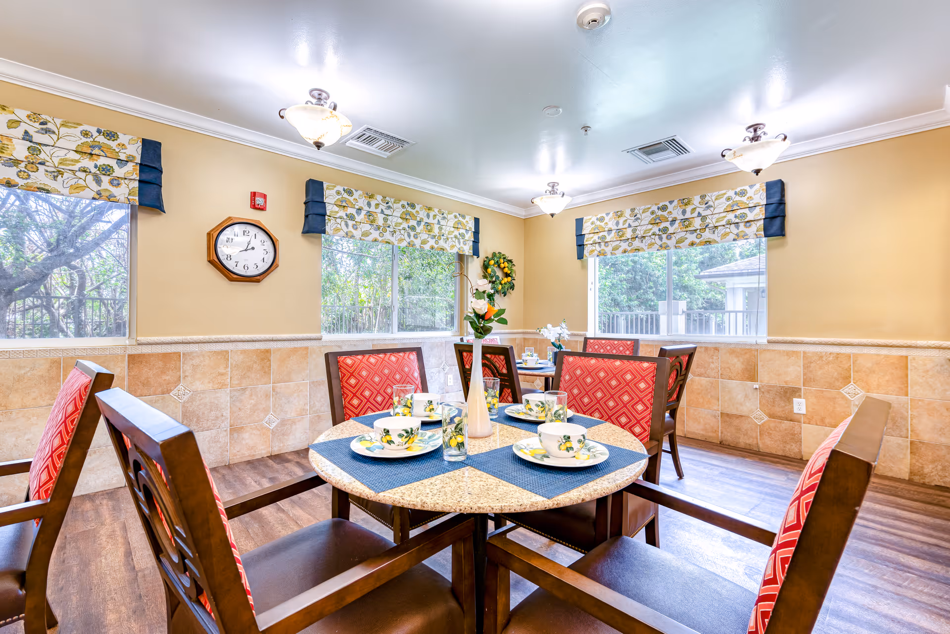 A bright dining room with round tables set for four people. Each table has blue placemats, floral-patterned dishes, and a vase with flowers. The chairs have wooden frames with red patterned cushions. The walls are painted beige with tiled wainscoting, and there are three windows with floral valances letting in natural light. A clock is mounted on one wall.