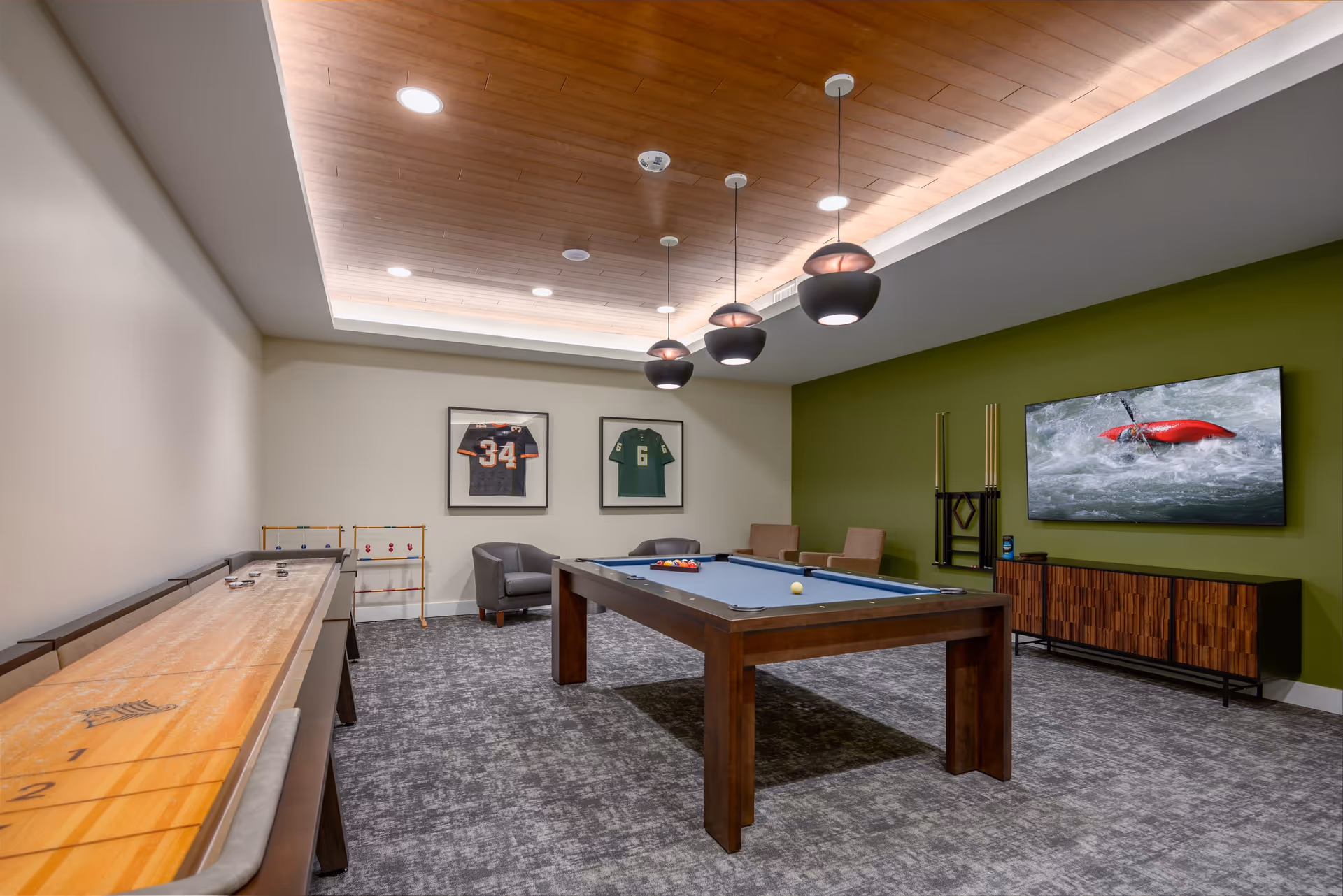 A recreational room featuring a pool table with balls and cues, a shuffleboard table, two framed sports jerseys on the wall, a flat-screen TV mounted on a green accent wall displaying a kayaking scene, and seating including two armchairs and a small table. The ceiling has wooden paneling with recessed lighting and hanging pendant lights.
