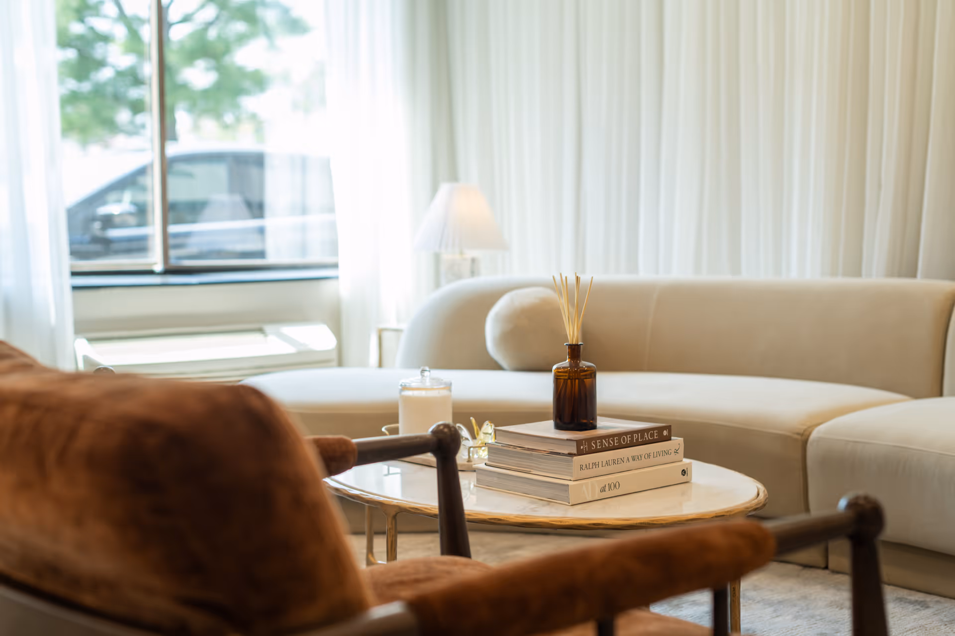 A cozy living room area with a beige curved sofa, a round coffee table with stacked books, a brown glass diffuser, and a candle. In the foreground, there is a brown cushioned chair. A window with sheer white curtains lets in natural light, and a table lamp is visible behind the sofa.