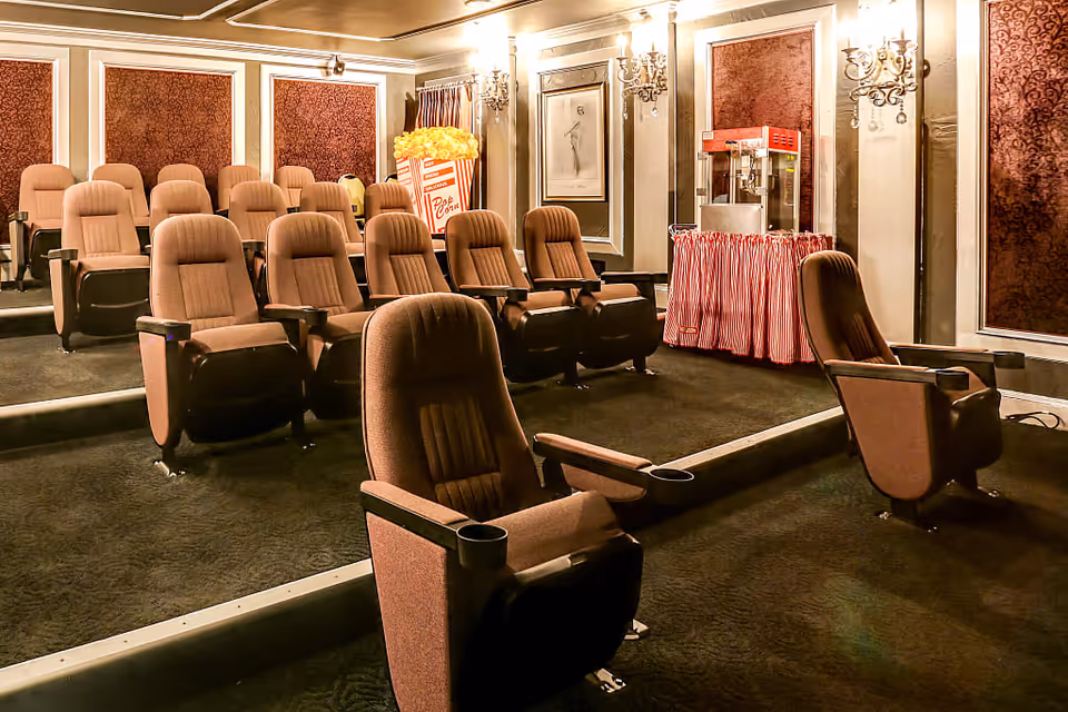 A small movie theater room with multiple rows of brown cushioned seats arranged in tiers. The room has ornate wall sconces with lights, decorative wall panels, and a popcorn machine with a large popcorn container in the corner.
