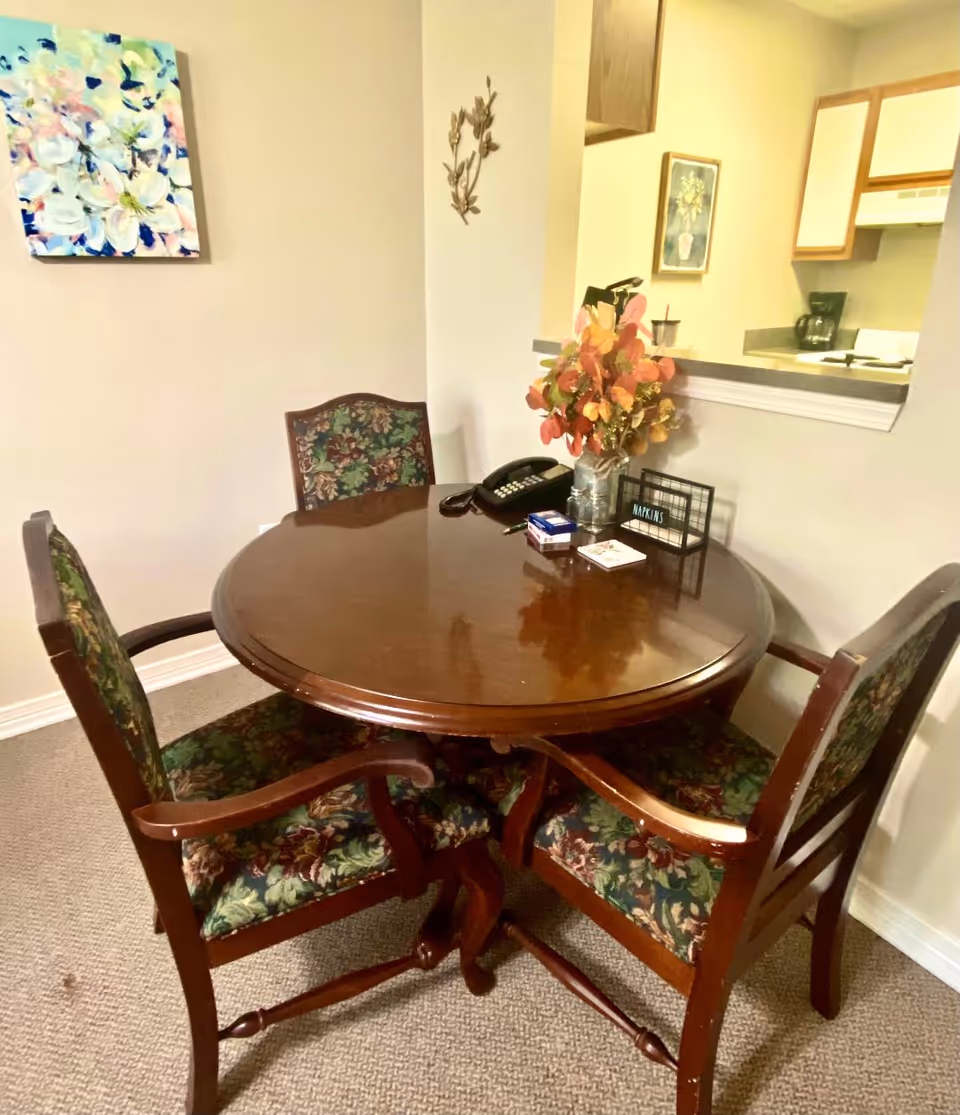 A round wooden dining table with four floral upholstered chairs around it. On the table are a vase with autumn-colored leaves, a telephone, a small sign labeled 'NAPKINS', and some small items. The table is set against a beige wall with a floral painting and a small metal wall decoration. A kitchen area with white cabinets and a coffee maker is visible through a pass-through window.