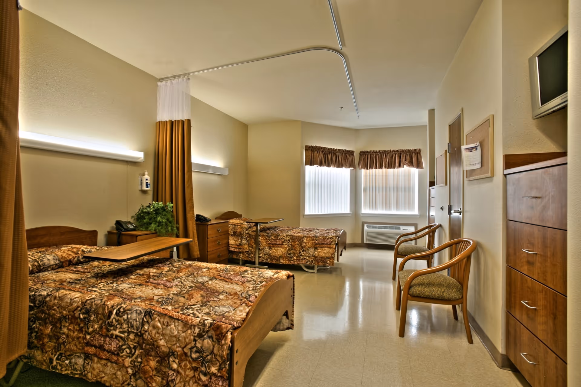 A well-lit senior care facility bedroom with two beds covered in patterned brown bedspreads. Each bed has a wooden headboard and an overbed table. There is a privacy curtain between the beds. The room has two windows with brown valances, a wall-mounted air conditioning unit, two wooden chairs with cushions, a wooden dresser, and a wall-mounted TV. The walls are light-colored, and the floor is a polished light tile.