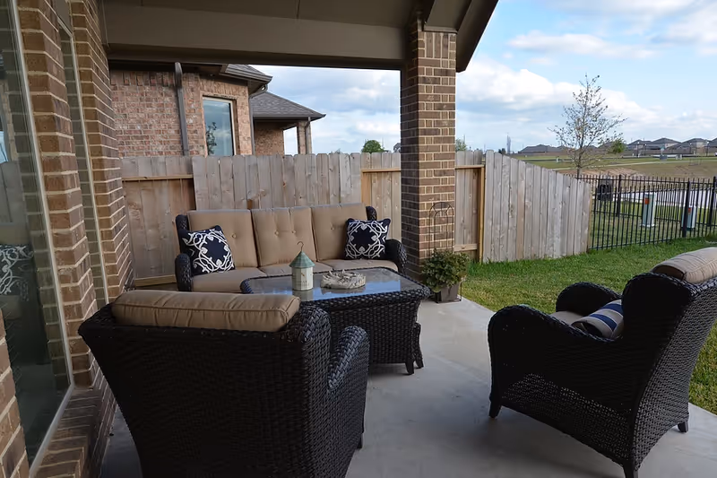 Covered backyard patio with wicker sofa and chairs arranged around a glass-top coffee table overlooking a fenced yard.