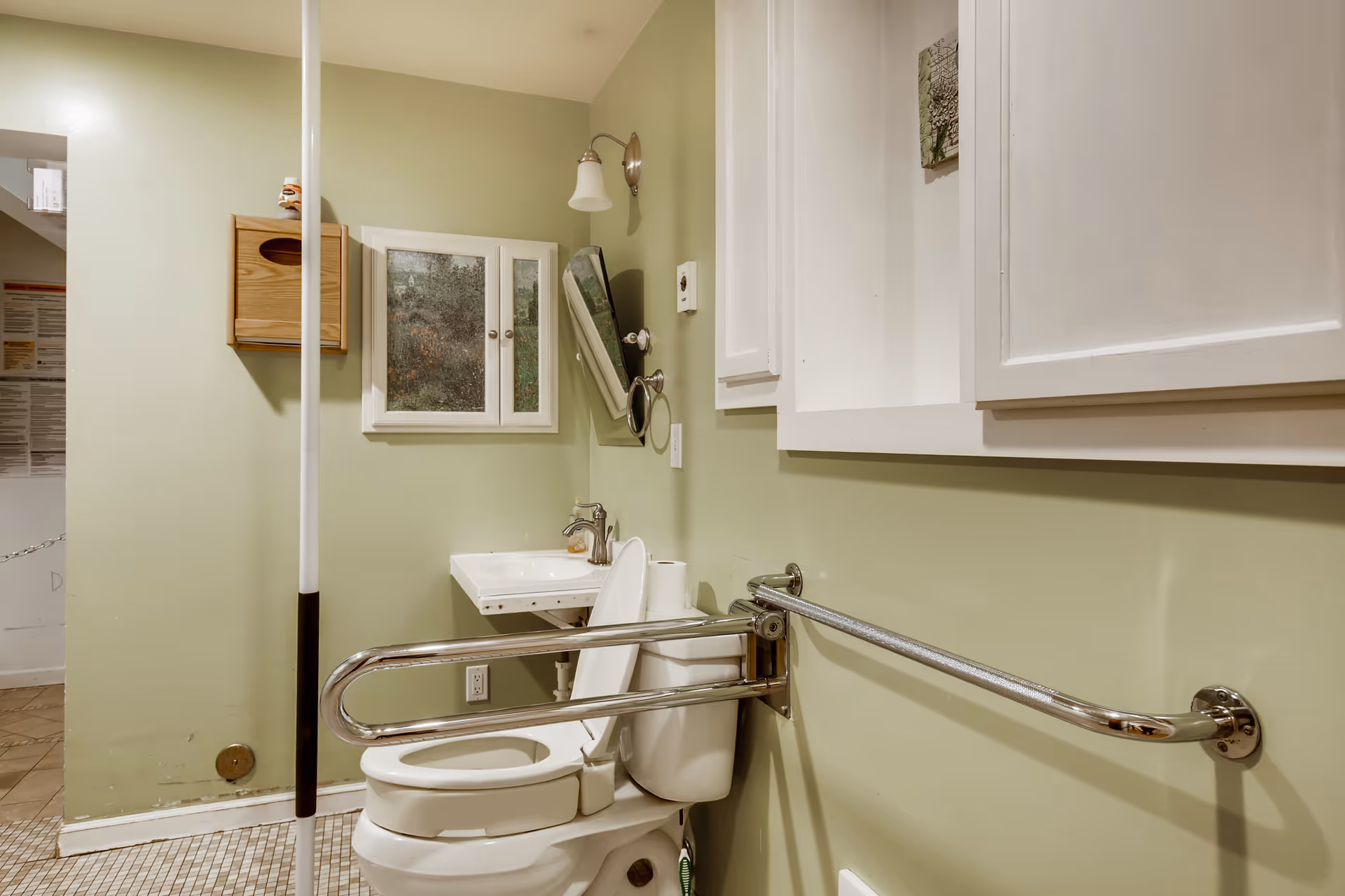 A bathroom with light green walls featuring a white toilet with the seat up and a metal grab bar installed on the wall beside it. There is a small white sink with a faucet and a wall-mounted mirror above it. A wooden tissue holder is mounted on the wall, and white cabinets are visible on the right side. The floor has small square tiles.