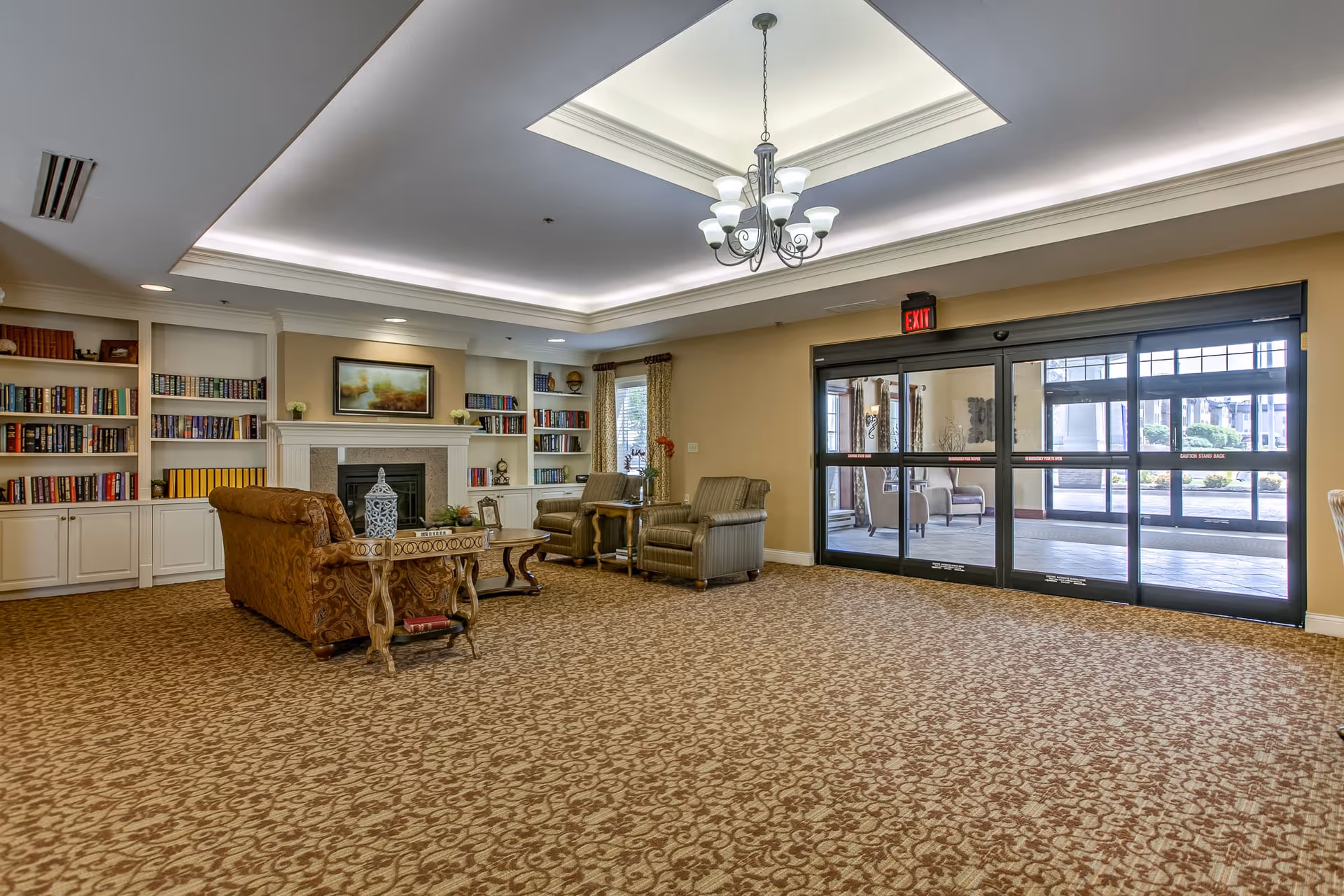A spacious and well-lit retirement community lobby with patterned carpet flooring, a chandelier hanging from a recessed ceiling, a seating area with a sofa and two armchairs around a coffee table, built-in bookshelves filled with books, a fireplace with a framed painting above it, and large glass automatic sliding doors leading outside.