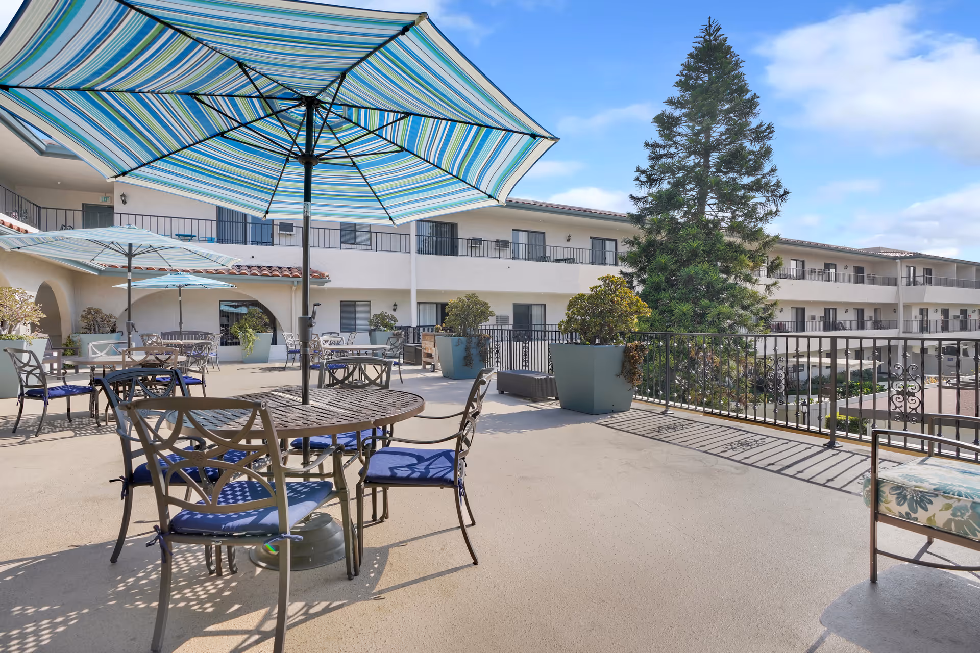 Outdoor terrace with metal tables and chairs under striped umbrellas, potted plants, and a multi-story residence in the background.