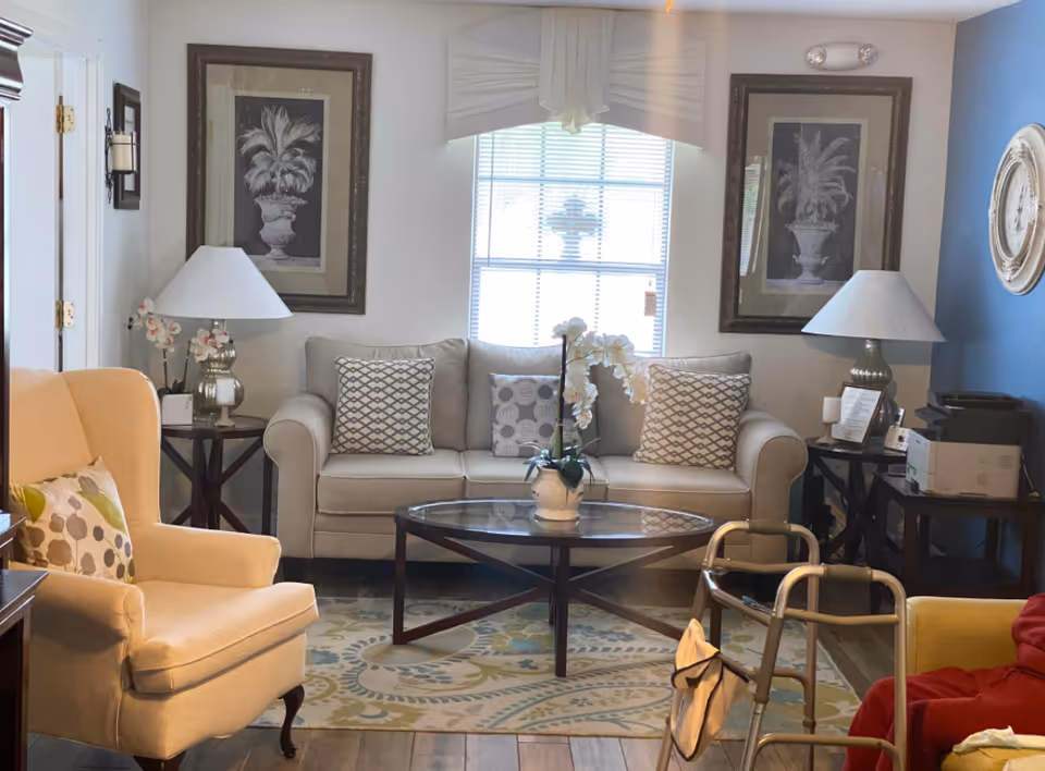 A cozy living room with a beige sofa adorned with patterned cushions, a glass coffee table with a white orchid centerpiece, two side tables each with a lamp, framed botanical artwork on the walls, a beige armchair, a walker, and a window with blinds letting in natural light.