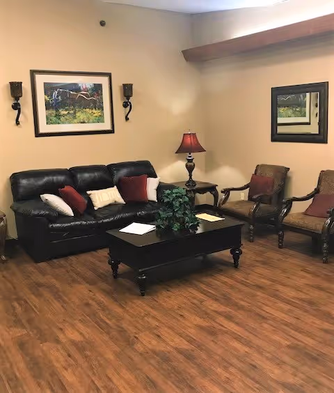 A cozy living room with a black leather sofa adorned with white and red pillows, two wooden armchairs with red cushions, a dark wooden coffee table with a green plant and papers on it, a side table with a red lamp, a framed painting on the wall, and a mirror reflecting part of the room. The floor is wooden and the walls are beige.