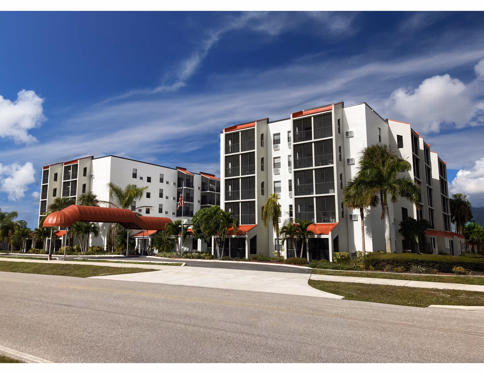 Exterior front of a multi-story white senior living building with red entrance canopy, screened balconies and palm trees under a blue sky.