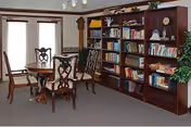 Interior common room with wooden bookshelves filled with books, a round wooden table and chairs, and windows with shades.