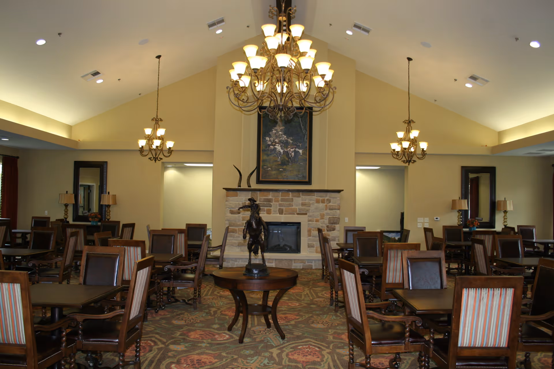 A spacious dining room with multiple wooden tables and chairs arranged neatly. The room features a high vaulted ceiling with three ornate chandeliers providing warm lighting. A stone fireplace with a painting above it is centered on the far wall, and a decorative statue is placed on a round table in front of the fireplace. The room has a patterned carpet and neutral-colored walls with mirrors and lamps on the side walls.