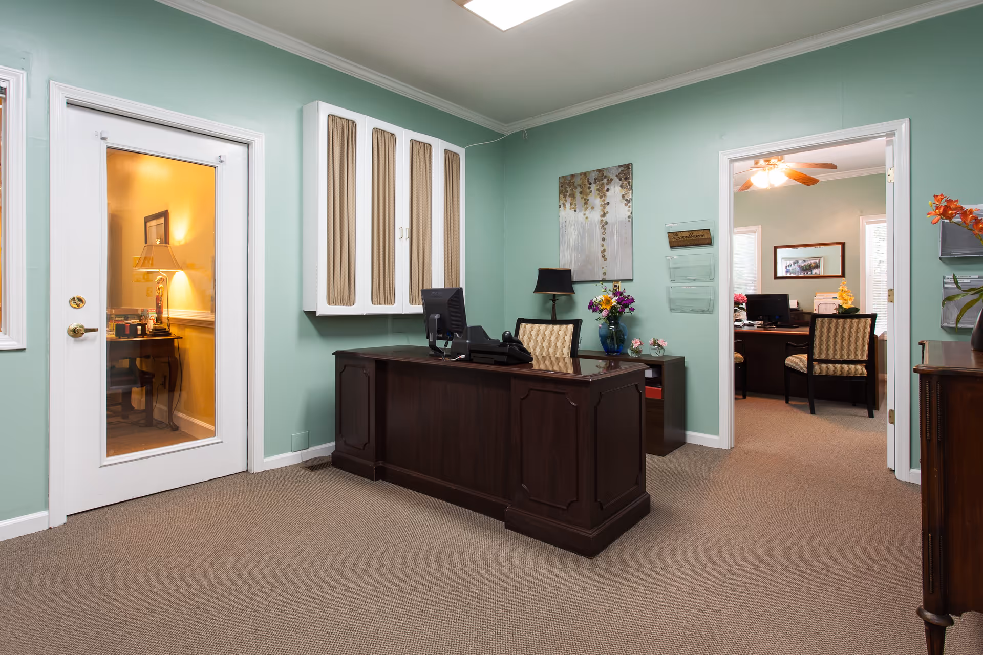 Reception area with a dark wooden desk, computer, and telephone. The walls are painted light green with a decorative painting and a lamp on a side table. There are two doorways, one with a glass door showing a warmly lit room with a lamp and framed picture, and another open doorway leading to an office with chairs and a ceiling fan.