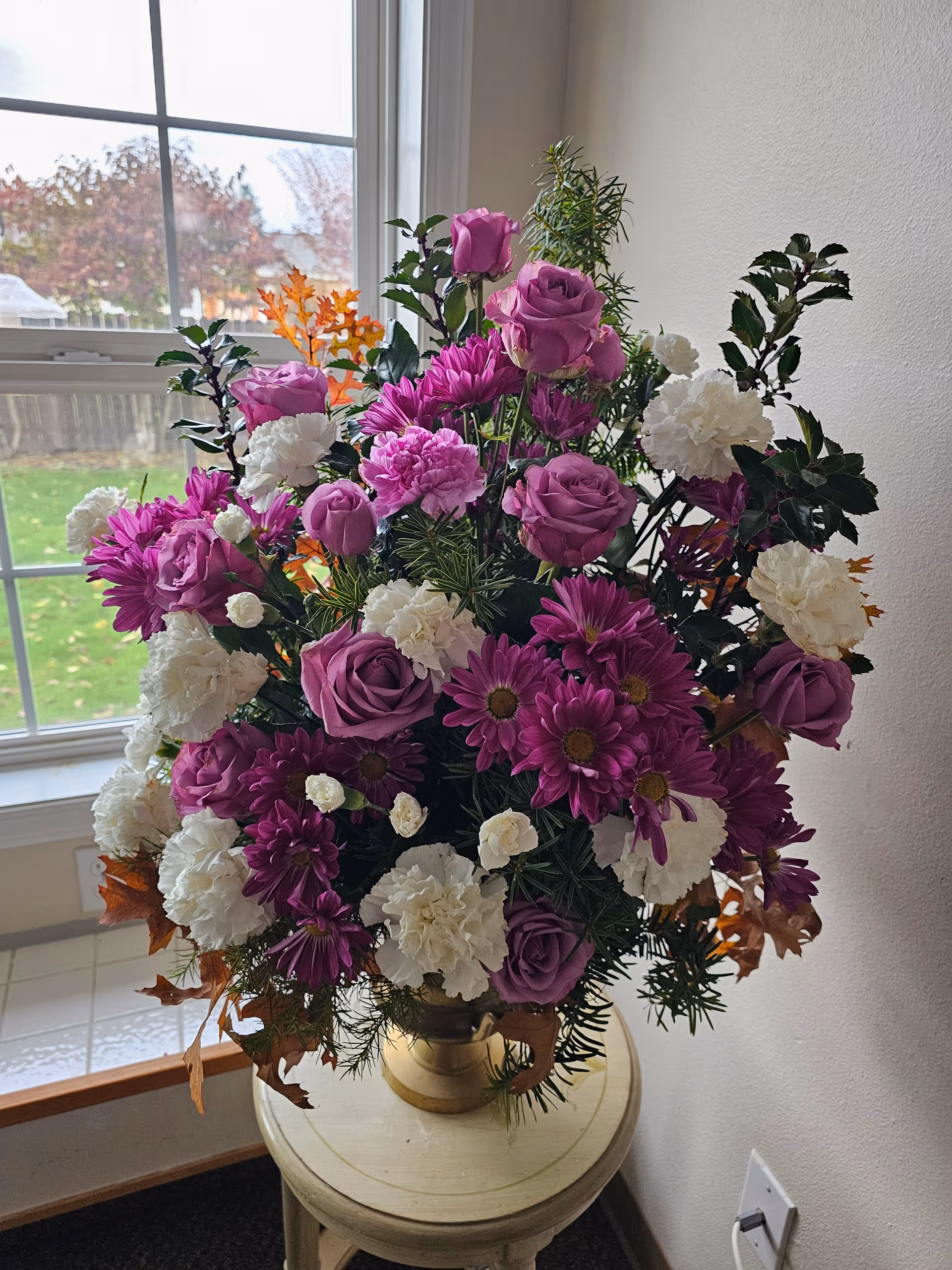 A large floral arrangement with purple roses, purple daisies, white carnations, and green foliage placed on a small round wooden table near a window with a view of a green lawn and trees outside.