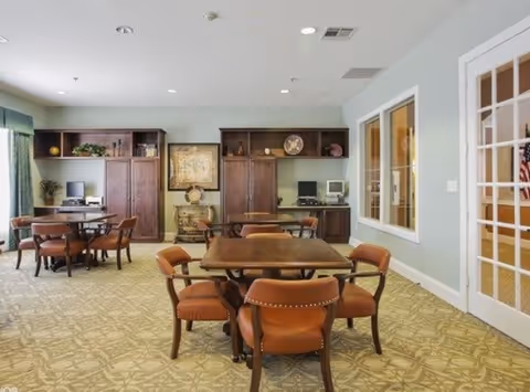 A well-lit room with multiple wooden tables and brown cushioned chairs arranged for seating. The room has a patterned carpet, light-colored walls, and built-in wooden cabinets along the back wall with decorative items and two computer workstations. There is a large window on the right wall and a glass door with white framing on the right side of the image.