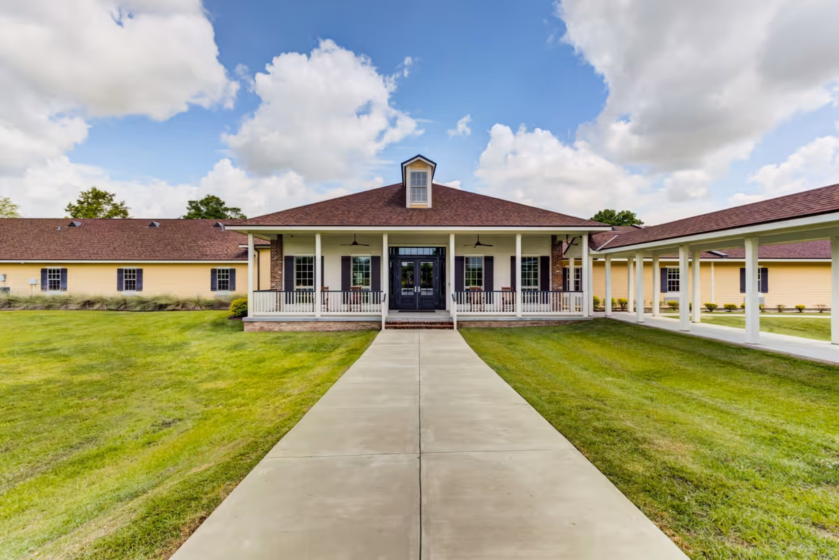 Front exterior view of a single-story building with a covered porch and a concrete walkway leading up to the entrance. The building has a brown roof, yellow siding, and white columns supporting the porch roof. There is a covered walkway extending from the right side of the building. The sky is partly cloudy.