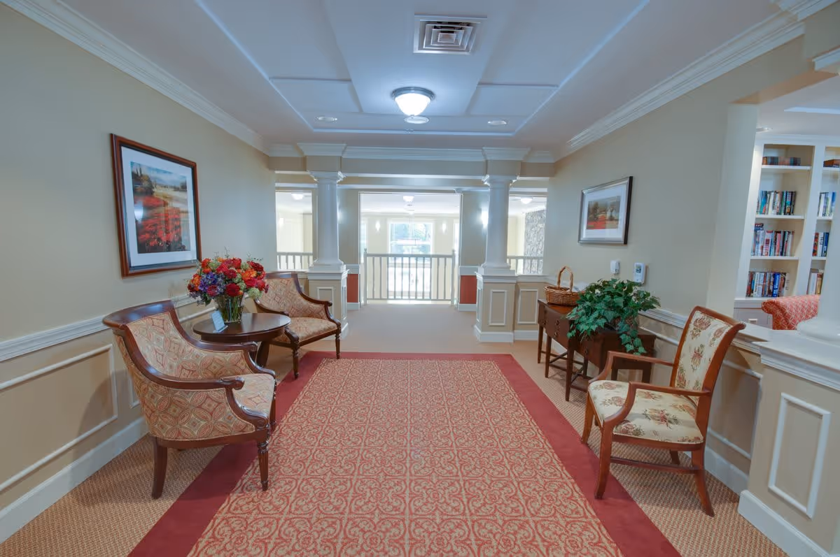 A well-lit hallway in a senior living facility with beige walls and white trim. The hallway features a patterned red carpet runner, two upholstered chairs with a small round table holding a flower arrangement on the left, and a wooden side table with a plant and basket on the right. There are framed pictures on the walls and a built-in bookshelf visible in an adjacent room.