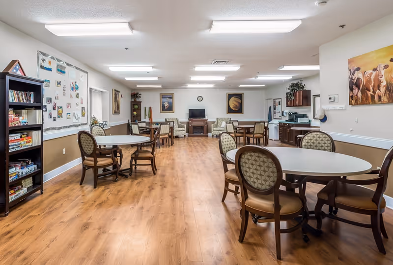 A spacious senior community activity room with round dining tables and chairs, bookshelves, a TV, kitchenette and wood-look flooring.