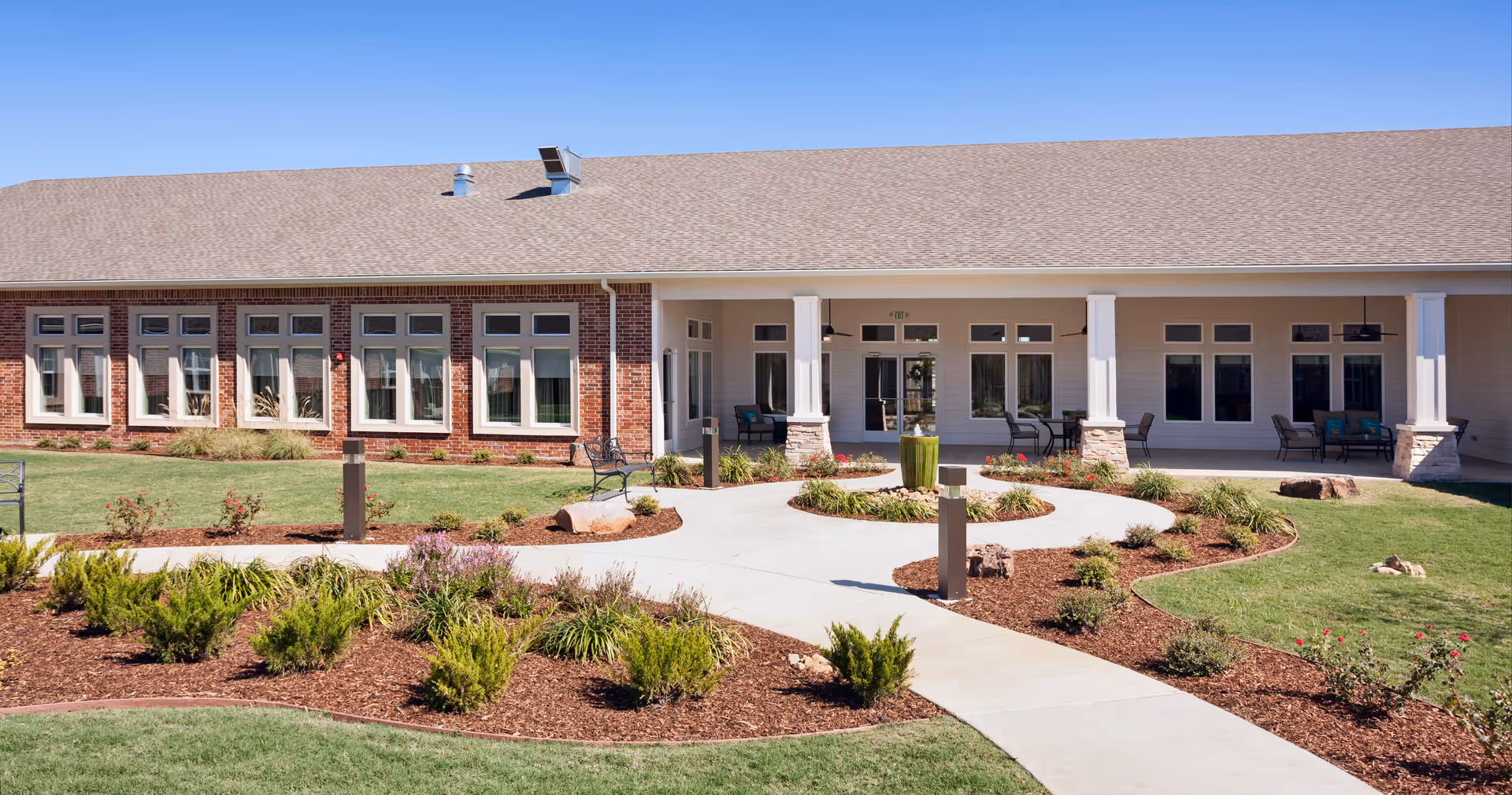 Exterior view of a single-story assisted living facility building with a brick and siding facade, multiple windows, a covered patio area with seating, and a landscaped garden with shrubs, flowers, and a paved walkway leading to the entrance.