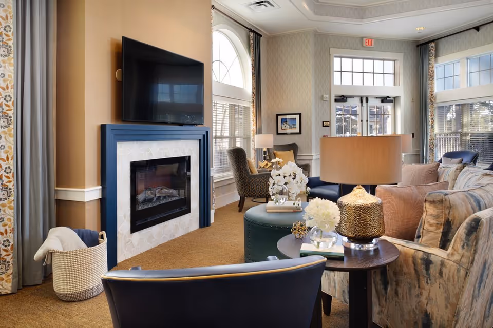 A cozy living room area with a mounted flat-screen TV above a modern fireplace. The room features comfortable seating including a patterned armchair, a blue chair, and a sofa with various cushions. There are decorative elements such as a table lamp with a textured gold base, white floral arrangements, and books on a round side table. Large windows with patterned curtains allow natural light to fill the space, and an exit door is visible in the background.