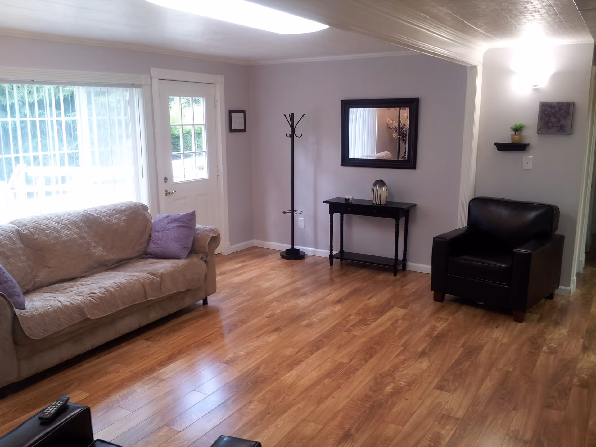 A living room with wooden flooring, a beige couch with purple pillows, a black armchair, a black coat rack, a small black table with a decorative item, a wall mirror, and a door with a window leading outside.