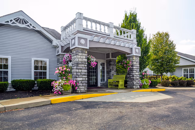 Covered front entrance of a senior living facility with stone pillars, hanging flower baskets, and a green bench.