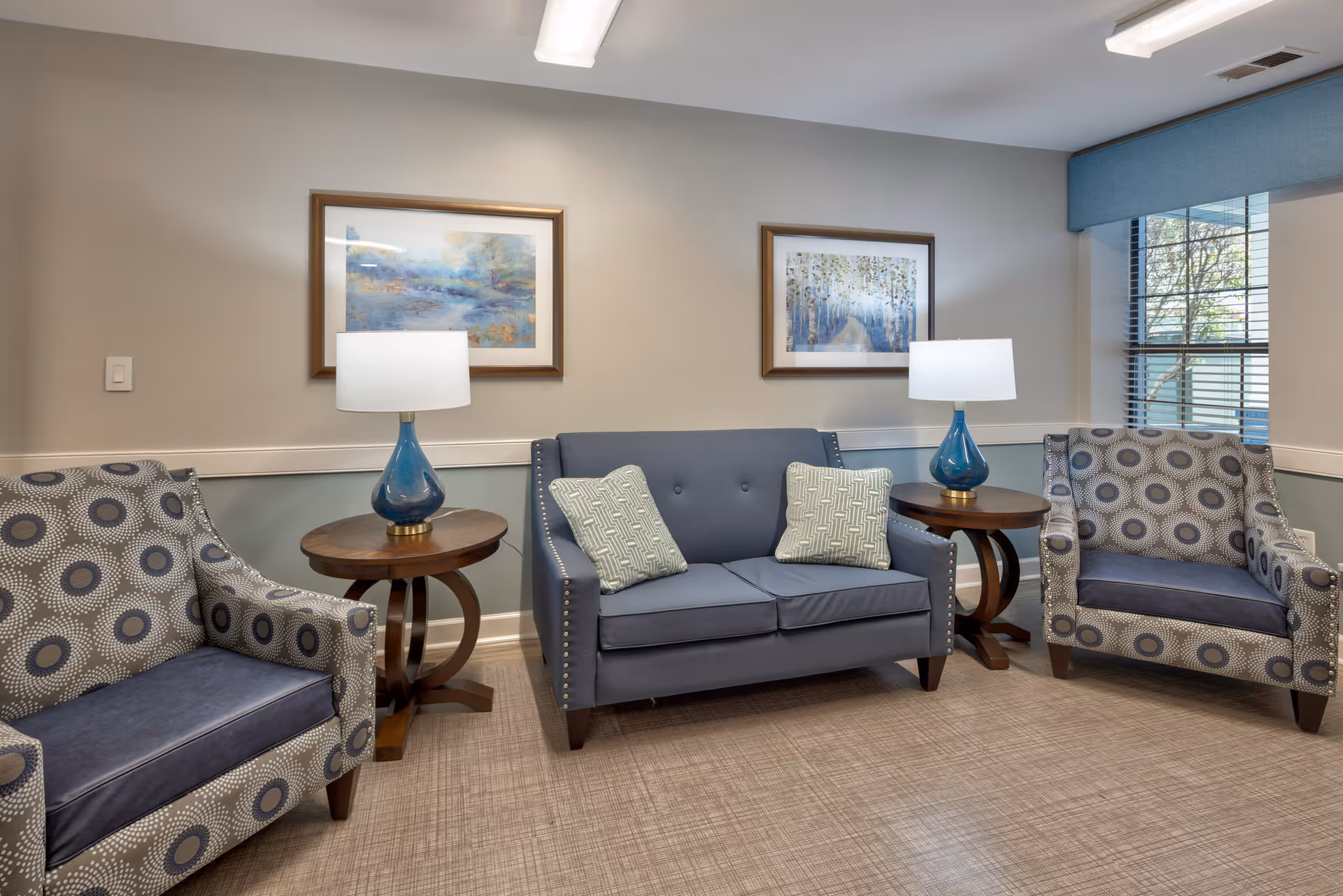 A cozy sitting area with a blue loveseat adorned with two patterned pillows, flanked by two patterned armchairs. Each side of the loveseat has a round wooden side table with a blue glass lamp. Two framed landscape paintings hang on the beige wall behind the loveseat, and a window with blinds is visible on the right side of the room.
