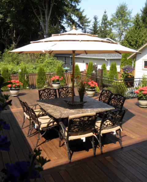 A wooden outdoor patio with a central table, umbrella, chairs, and potted plants.