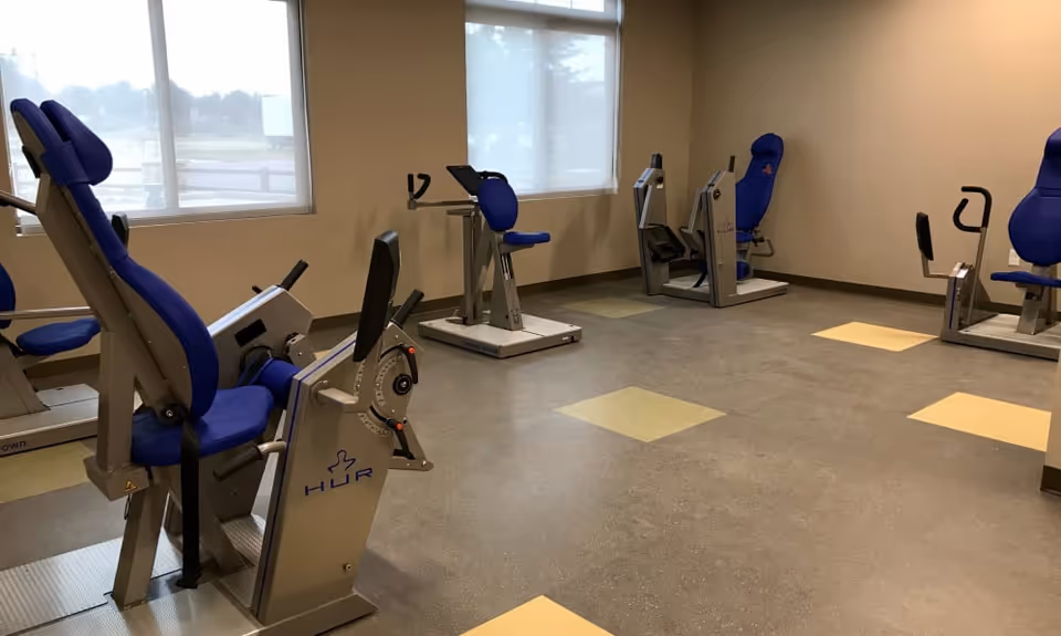 A fitness room with several exercise machines featuring blue padded seats and backrests, positioned near windows with white blinds. The floor has a speckled pattern with square colored tiles in yellow and green shades.