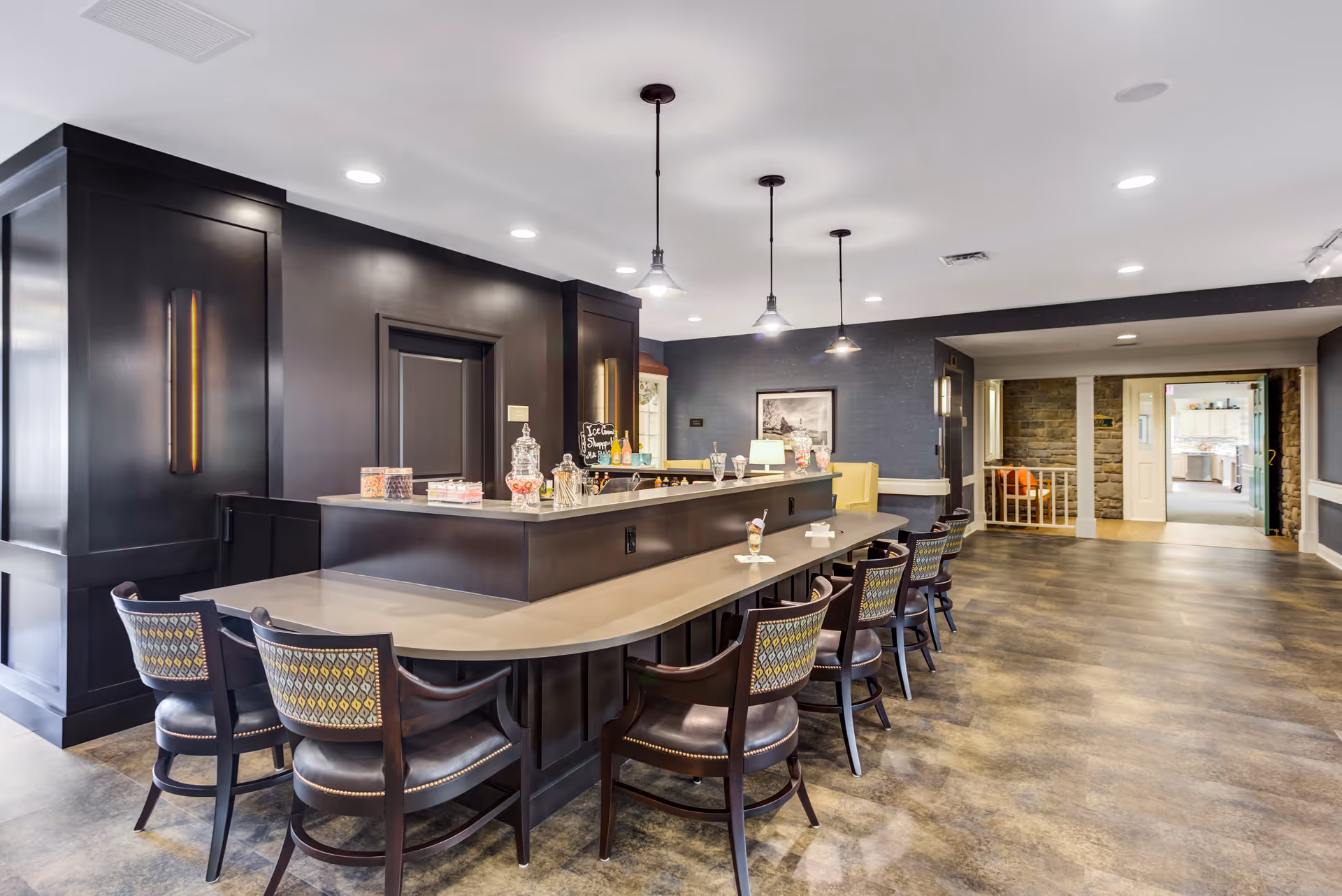Interior view of a senior living facility featuring a long counter with several chairs arranged along it. The counter has jars of candy and ice cream sundae glasses on it. The room has dark wood paneling, pendant lights hanging from the ceiling, and a seating area with a yellow armchair in the background. The floor is covered with wood-patterned flooring and there is a doorway leading to another room with stone walls.