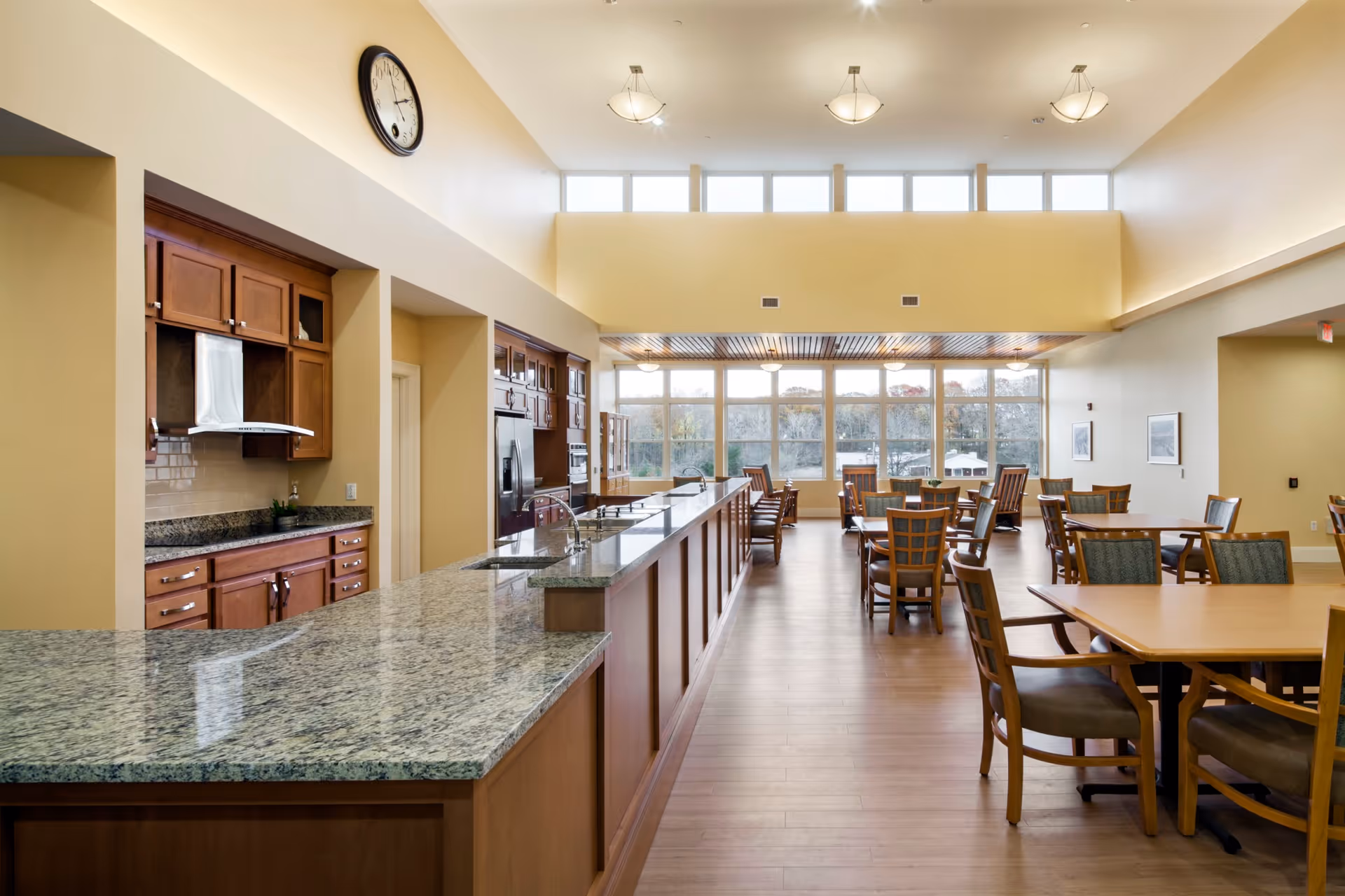 A spacious dining area with wooden tables and chairs arranged neatly. To the left is a large kitchen counter with granite countertops and wooden cabinets. The room has high ceilings with multiple light fixtures and large windows at the far end letting in natural light.