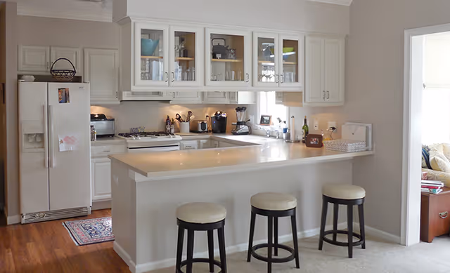 Bright open kitchen with white cabinets, a long breakfast bar with three stools, refrigerator, stove, and small countertop appliances.