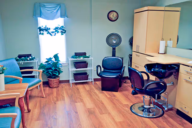 A salon room with wooden flooring featuring two green cushioned chairs with wooden arms, a small wooden table, a potted plant, a window with a blue valance and decorative greenery, a black salon chair in front of a hair washing station with a sink, a black dryer chair, a wall clock, and light wood cabinetry with a large mirror.