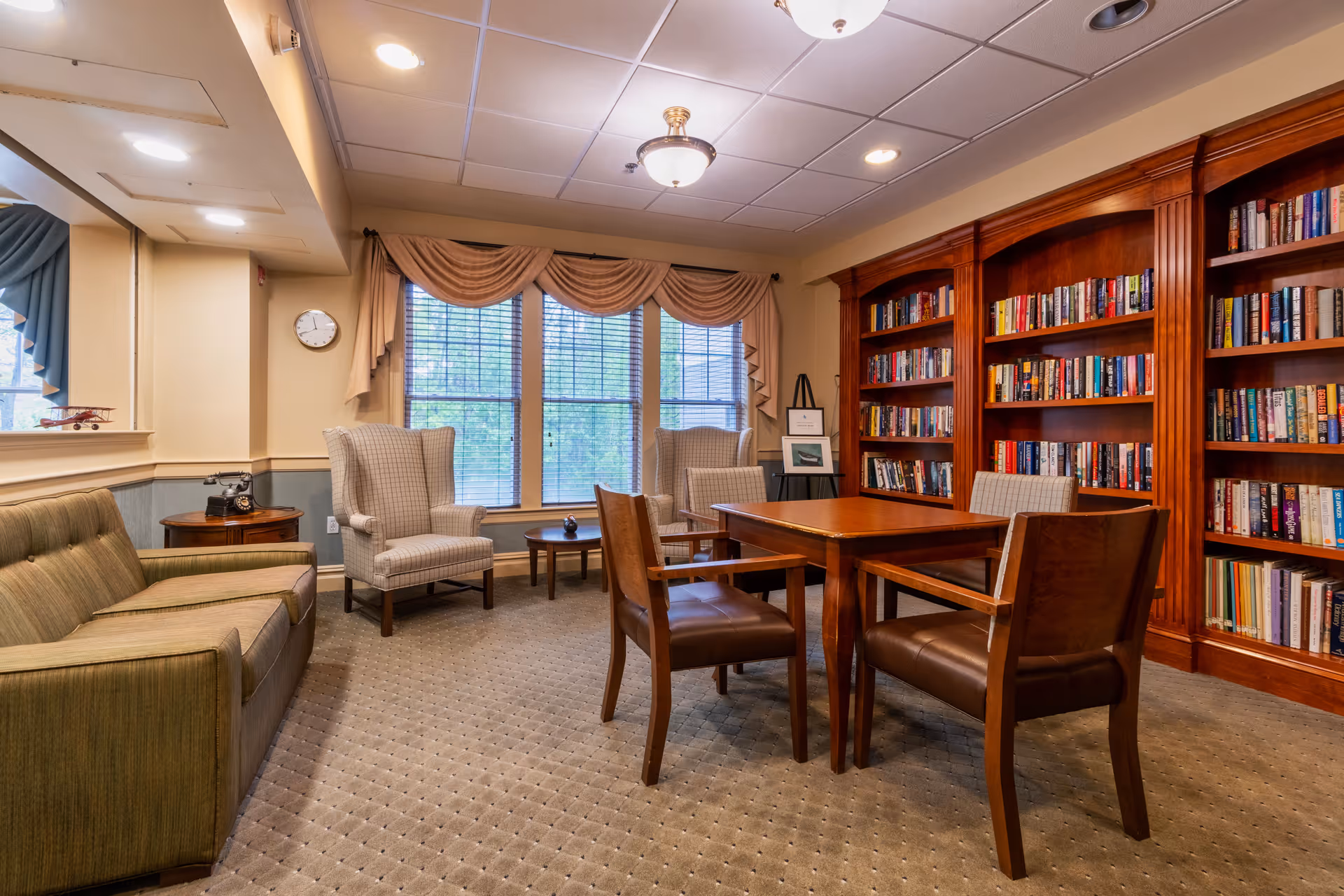 Cozy senior living common room with bookshelves, a table and chairs, armchairs and a sofa in front of large windows.