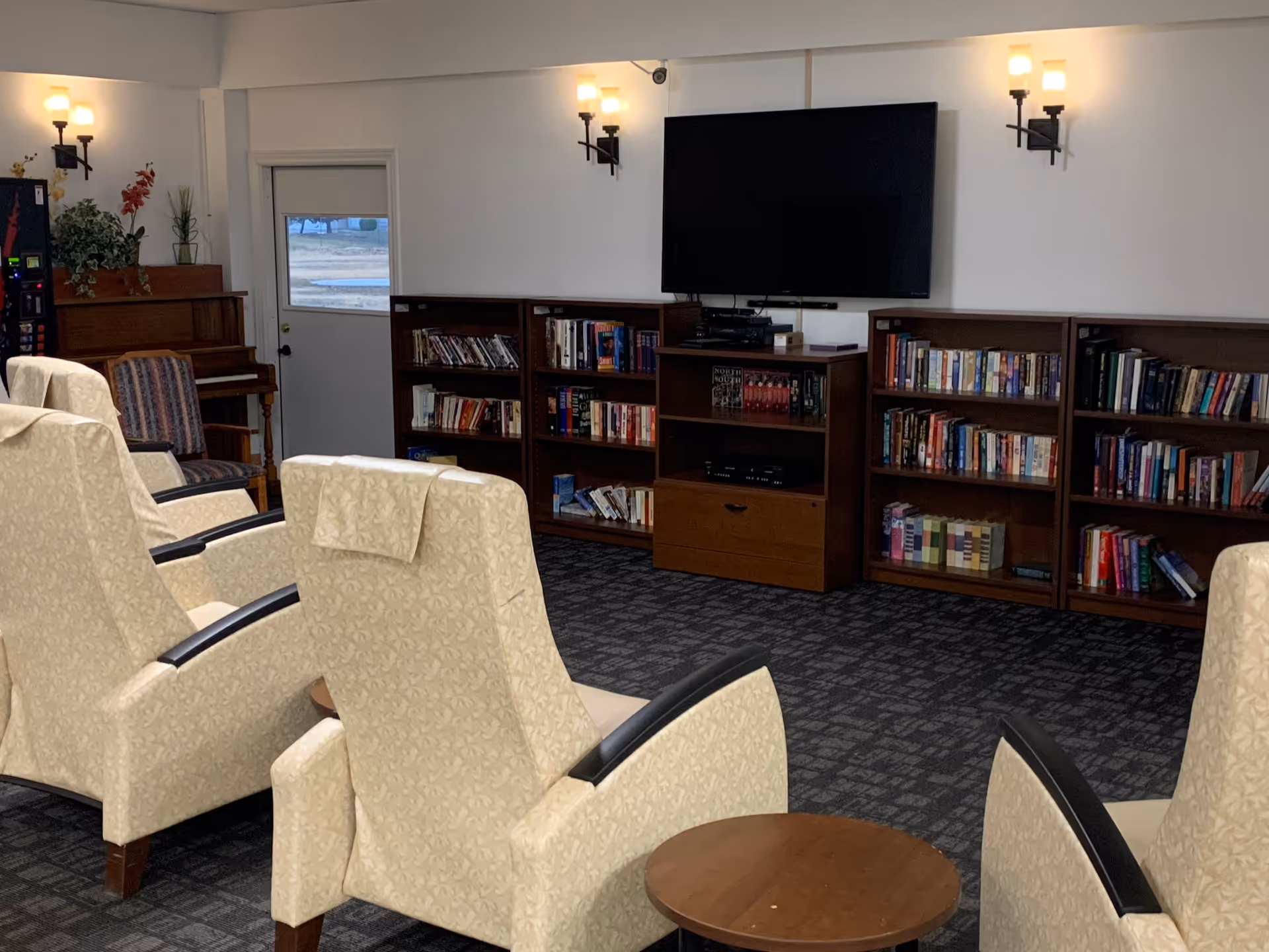A cozy common area with several beige upholstered armchairs arranged facing a wall-mounted flat-screen TV. Below the TV are wooden bookshelves filled with books and media equipment. To the left, there is a piano and a vending machine near a door with a window. The room has wall-mounted lights and a carpeted floor.