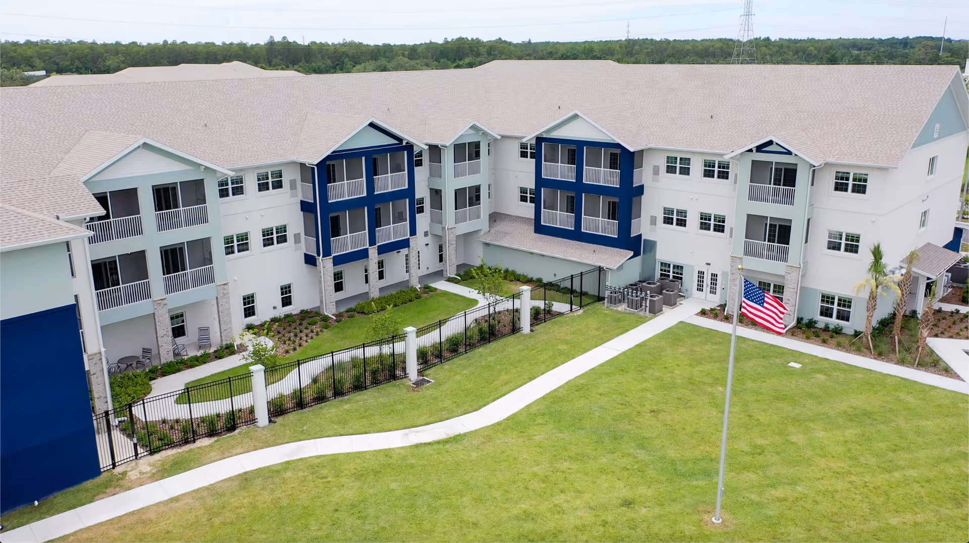 Exterior view of a multi-story senior living facility building with white and blue walls, balconies, a fenced garden area, a green lawn, and an American flag on a flagpole in front.