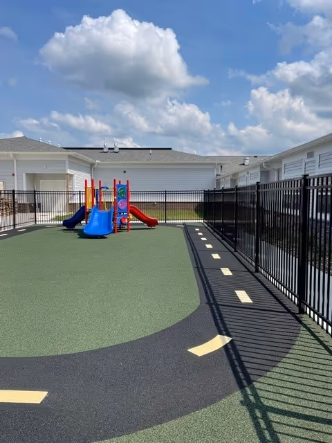 Outdoor fenced play area with a small colorful playground structure featuring blue and red slides, surrounded by a green and black rubberized surface with yellow markings, under a partly cloudy sky.
