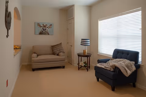 A cozy sitting area in a senior living facility with a beige loveseat adorned with a patterned pillow, a small round wooden side table with a lamp featuring a blue and white striped shade, and a dark blue armchair draped with a light-colored throw blanket. A large window with white blinds allows natural light to fill the room. A painting of a cow hangs on the wall above the loveseat.