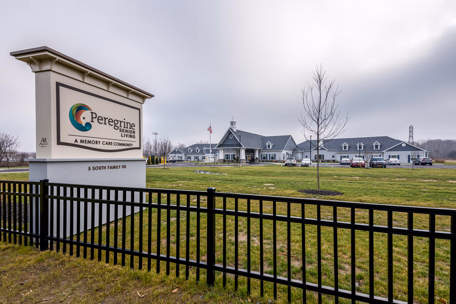 Exterior view of Peregrine Senior Living at Colonie, showing a large sign with the facility's name and logo in the foreground, a black metal fence, a grassy area, and the senior living building with parked cars in the background under a cloudy sky.
