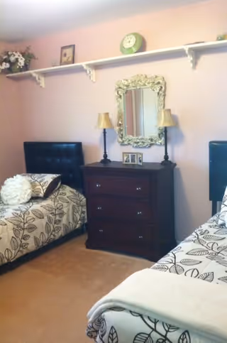 A cozy bedroom with two twin beds featuring black tufted headboards and matching leaf-patterned bedding. Between the beds is a dark wooden dresser with three drawers, topped with two lamps and a decorative ornate mirror hanging above it. A white shelf runs along the wall above the mirror, holding a green clock, framed pictures, and a flower arrangement. The walls are painted light pink and the floor is carpeted.