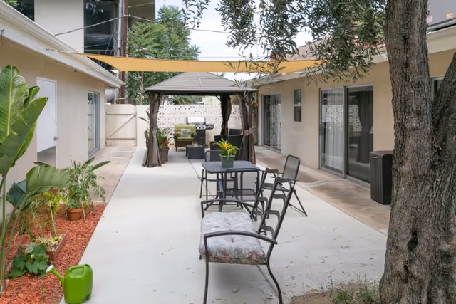 Outdoor courtyard area with a concrete pathway, metal chairs and tables, a bench with floral cushions, potted plants, a large tree, and a shaded gazebo with seating and a grill in the background.