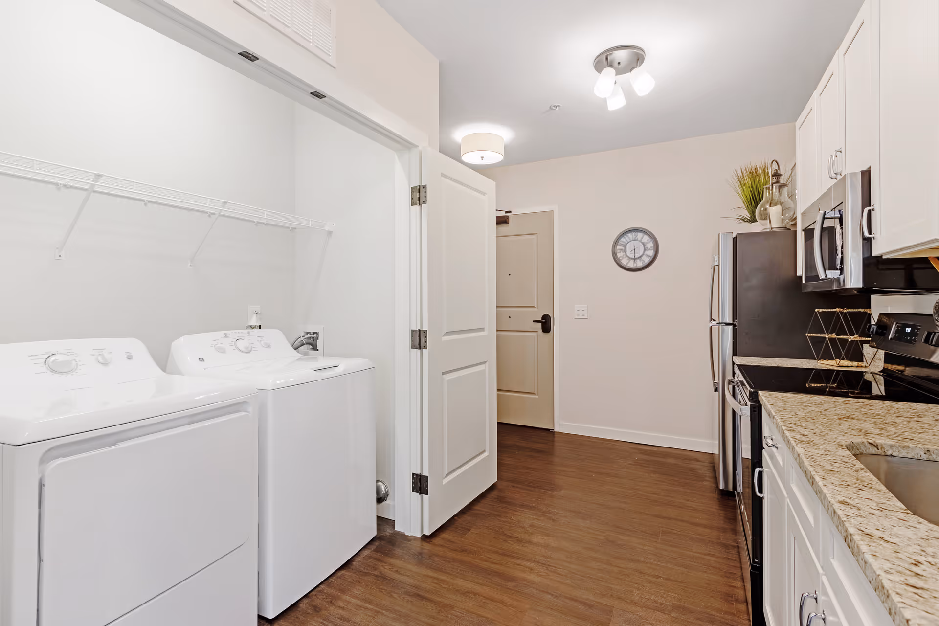 Interior view of a modern apartment kitchen with white cabinets, granite countertops, stainless steel appliances including a refrigerator, microwave, and stove. To the left, there is an open laundry area with a white washing machine and dryer under a wire shelf. The floor is wooden, and a clock is mounted on the wall near the entrance door.