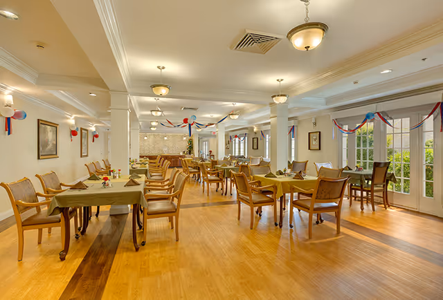 A spacious dining room with multiple tables covered in green tablecloths, each surrounded by wooden chairs with cushioned seats. The room is decorated with red, white, and blue streamers and balloons. Large windows and glass doors let in natural light, and ceiling lights provide additional illumination. The floor is wooden, and the walls are adorned with framed artwork.
