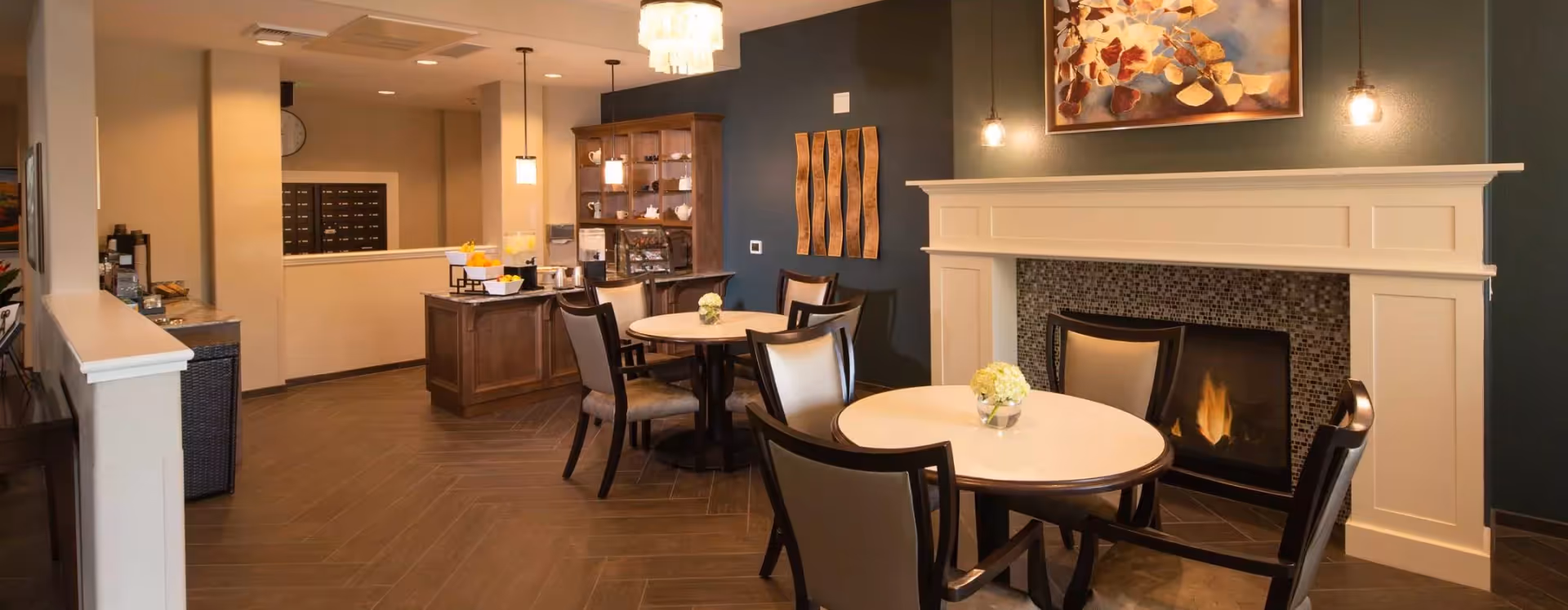 A cozy dining area with round tables and chairs arranged near a fireplace with a lit fire. The walls are painted in dark and light tones, decorated with artwork and wall hangings. Pendant lights hang from the ceiling, and there is a wooden cabinet and counter area in the background with various items on display.