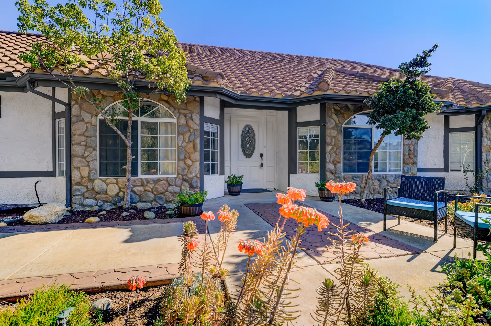 Front exterior view of a single-story building with a tiled roof, stone and stucco walls, and a white front door with an oval glass panel. There are two small trees and potted plants near the entrance, along with outdoor seating featuring black wicker chairs with blue cushions. Bright orange flowers and green shrubs are in the foreground under a clear blue sky.