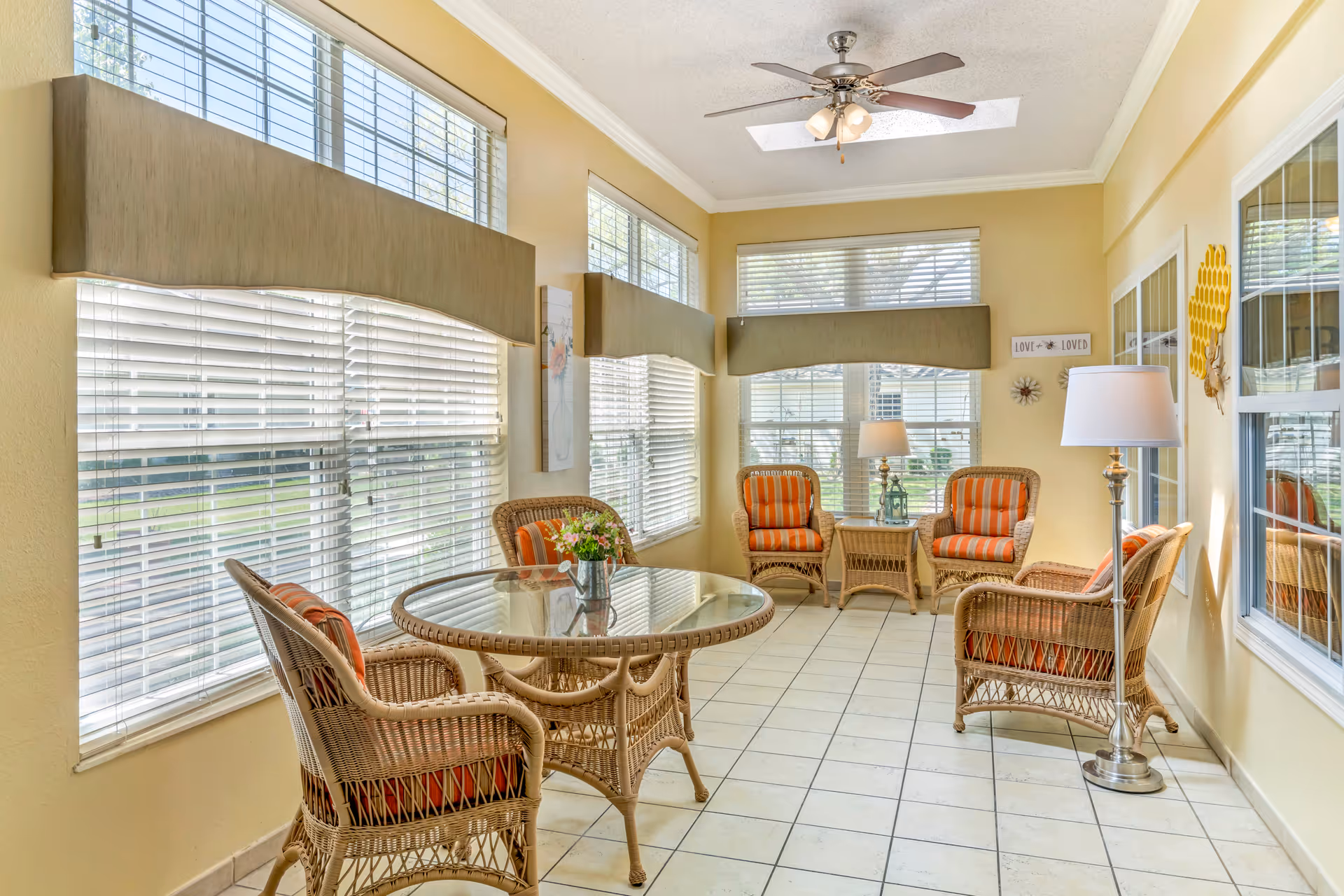 Bright sunroom with wicker chairs around a glass-top table, tiled floor, and large windows with blinds.